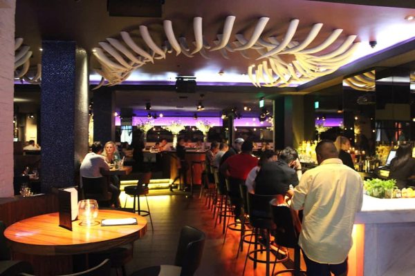 A dark bar and restaurant with people seated at tables and a long bar, illuminated by purple lights.