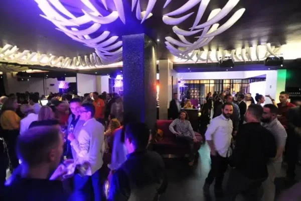 A crowded party in a dimly lit room with purple and blue lighting and white curved ceiling decorations.