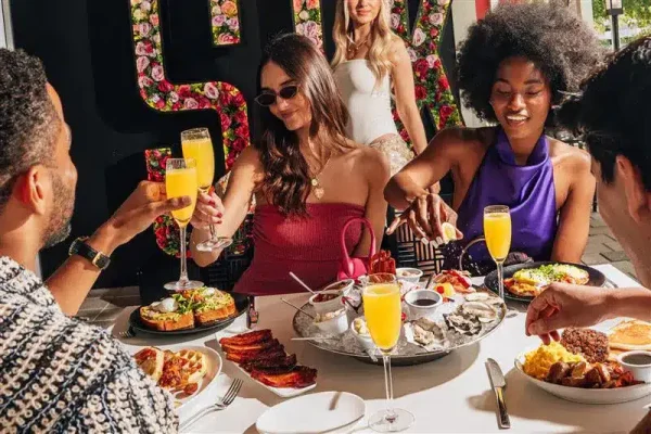 Four people enjoy brunch with mimosas, oysters, and other dishes at a table with a floral "SUMMER" sign.