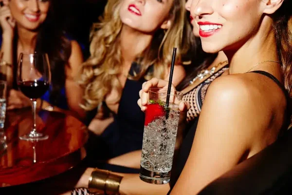 Three women are at a bar, one holding a tall glass with ice, a strawberry, and a black straw.